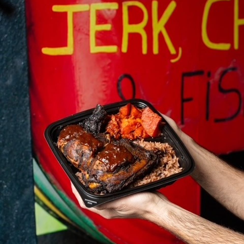 Hands hold a takeout tray filled with jerk chicken, rice, roasted plantains, and vegetables against a colorful restaurant mural.