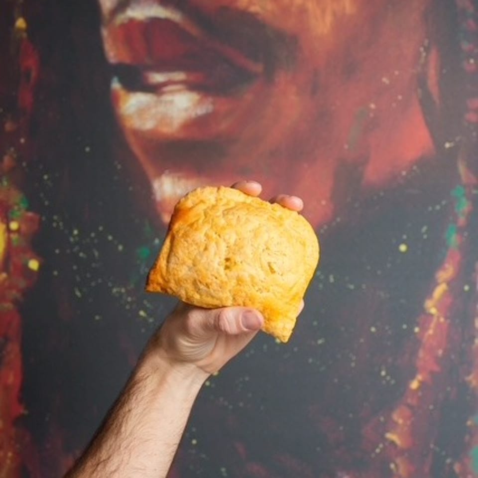 Hand holding a golden baked pastry in front of a colorful close-up portrait mural.