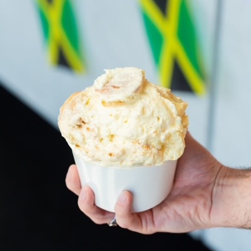 Hand holding a cup of creamy ice cream topped with a scoop, with Jamaican flags blurred in the background.